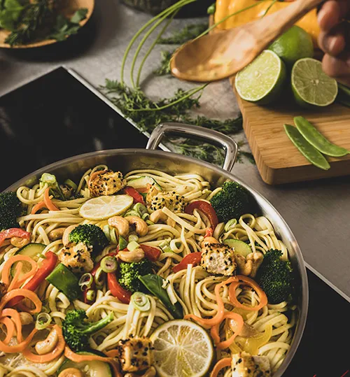 Asiatische Nudel Wok Rezept mit frischem Gemüse Wok mit Nudeln, Gemüse und Fleisch, daneben Limetten und Kräuter auf Holzbrett