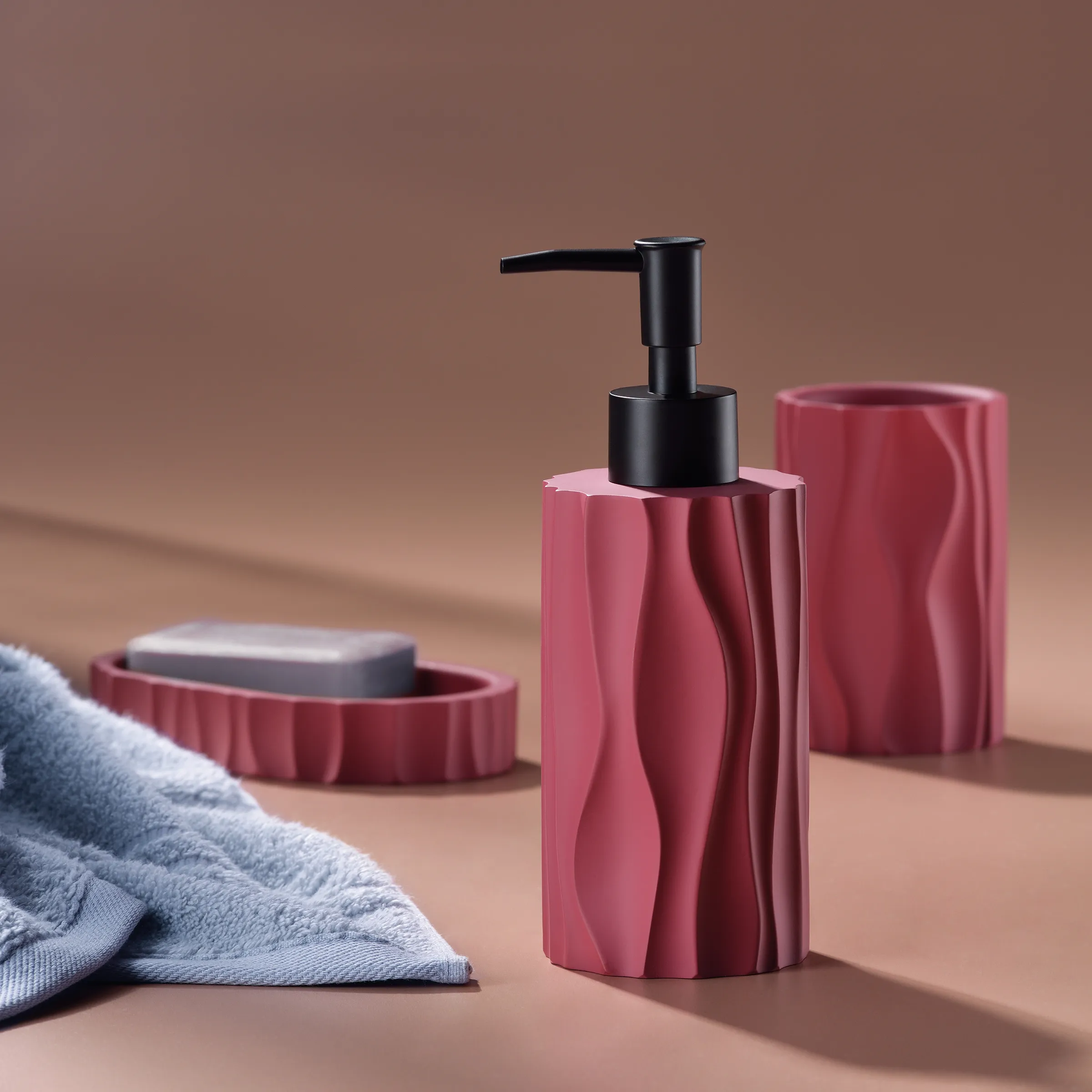 Rosa Badezimmer-Set mit strukturierter Oberfläche: Seifenspender mit schwarzem Pumpenkopf, Zahnputzbecher und Seifenschale neben grauem Handtuch