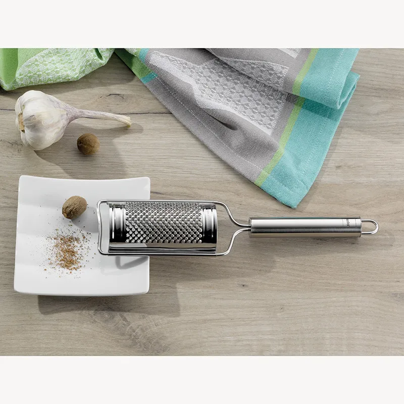 Muskatnussreibe Edelstahl - Küchenreibe Set Edelstahlreibe auf weißem Tablett mit geriebener Muskatnuss, daneben Muskatnussknollen und bunte Küchentücher auf Holztisch