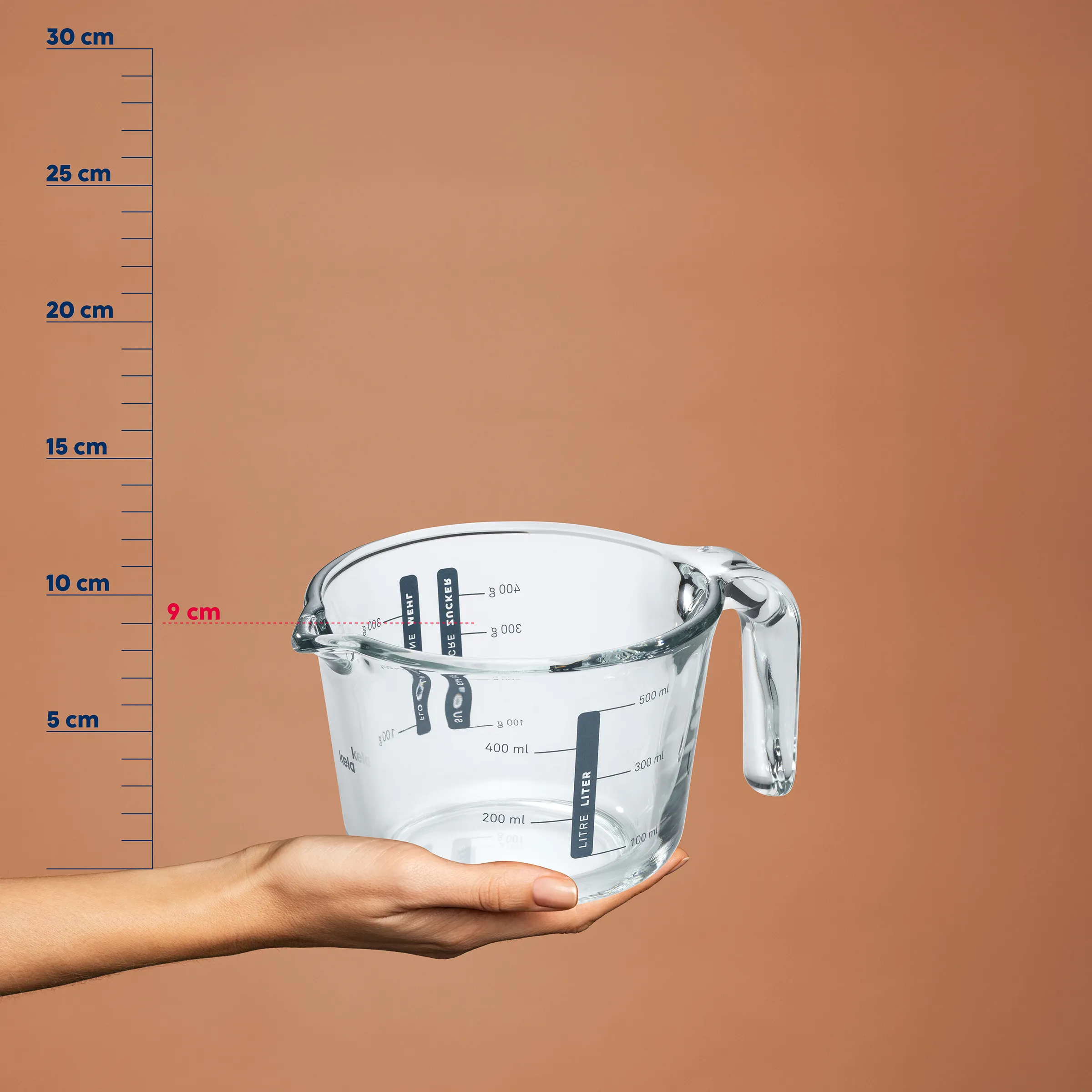 Litermaß Messbecher Glas 600ml mit Skala und Griff - Küchenmessbecher transparent Hand hält einen transparenten Litermaß-Messbecher mit Skala bis 600 ml vor einer braunen Größenskala