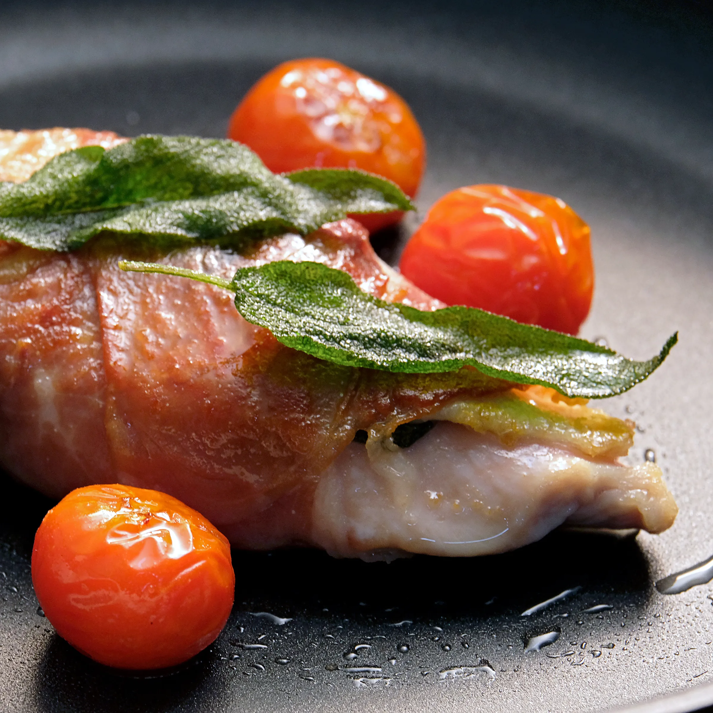 Speckumwickeltes Fischfilet mit Salbei und Kirschtomaten – Gourmet Fischgericht Speckumwickeltes Fischfilet mit Salbeiblättern und gebratenen Kirschtomaten auf dunklem Teller