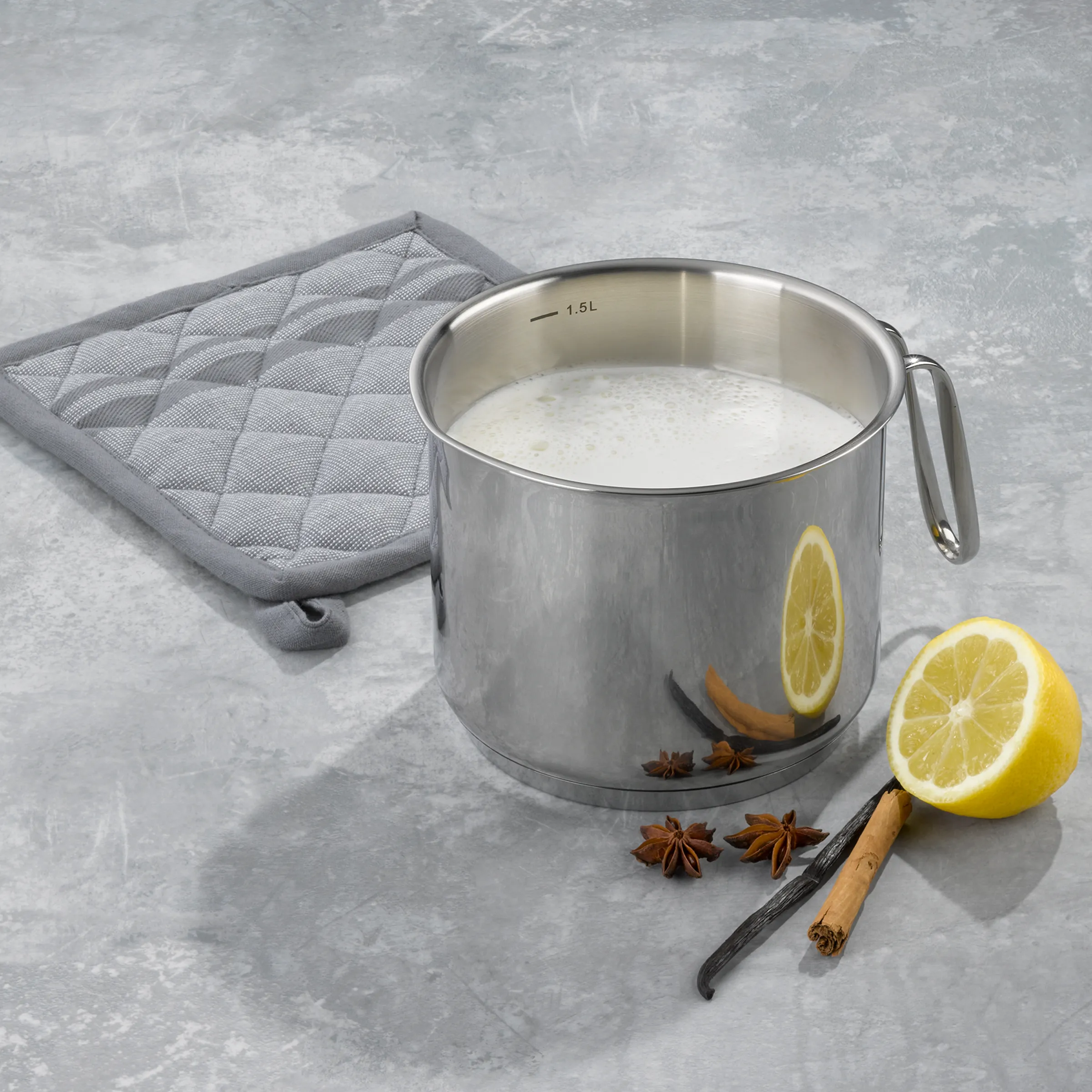 Edelstahl-Milchtopf 1,5 Liter mit Gewürzen - Kochtopf für Milch und Getränke Edelstahl-Milchtopf mit 1,5L Milch, Zitrone, Zimt, Sternanis und Vanille auf grauem Topflappen
