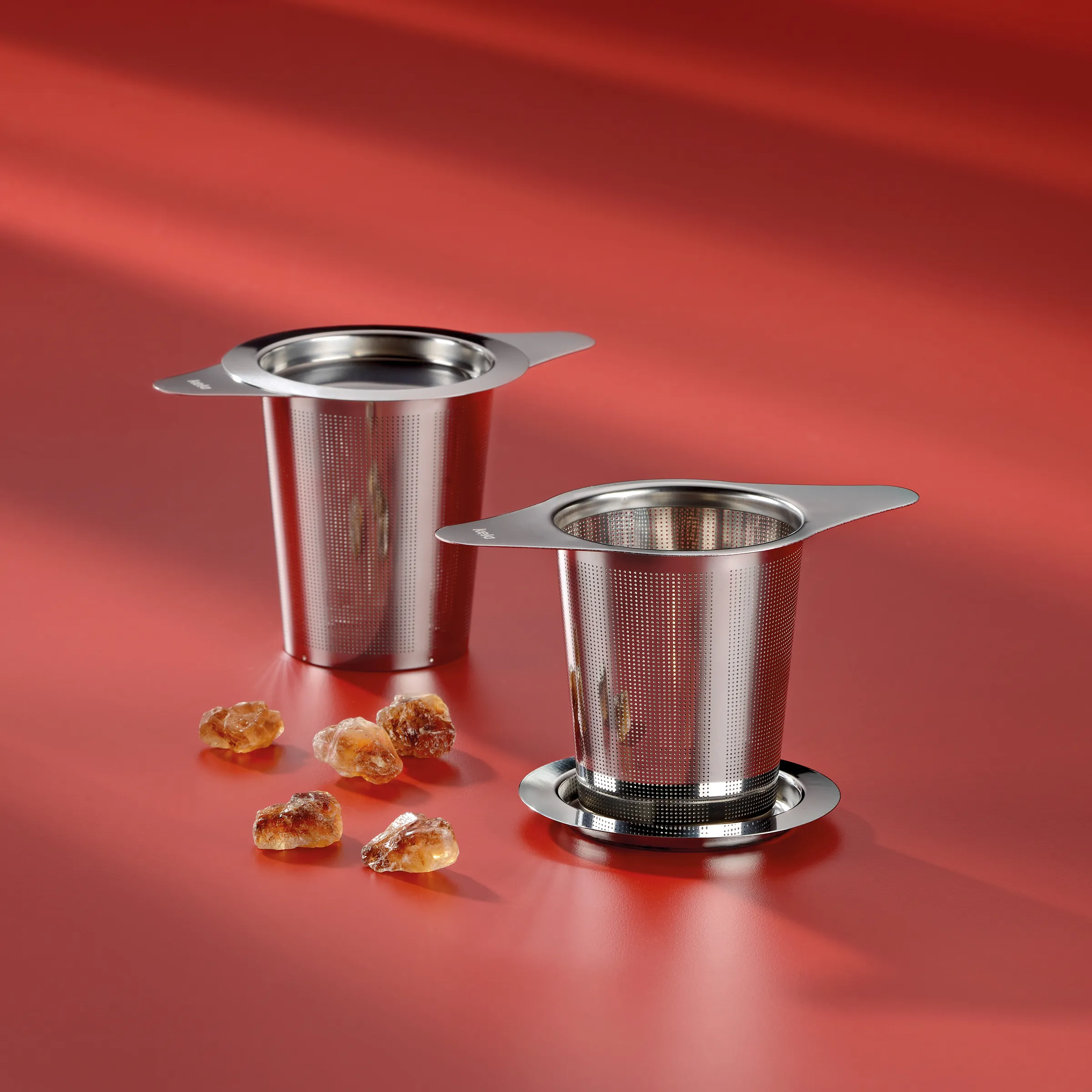 Zwei Edelstahl-Teeinfuser mit Griffen und Netzsieb auf rotem Hintergrund, daneben Kandiszucker-Kristalle