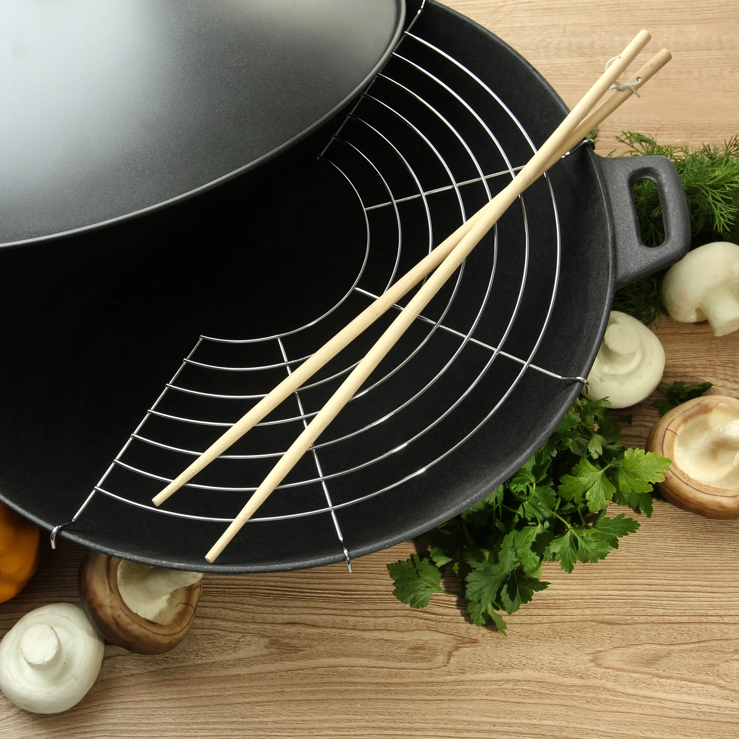 Wok-Pfanne mit Stäbchen und frischen Zutaten zum Kochen Schwarze Wok-Pfanne mit Holzstäbchen, umgeben von frischen Zutaten wie Zwiebeln, Kräutern und Gewürzen auf Holztisch.