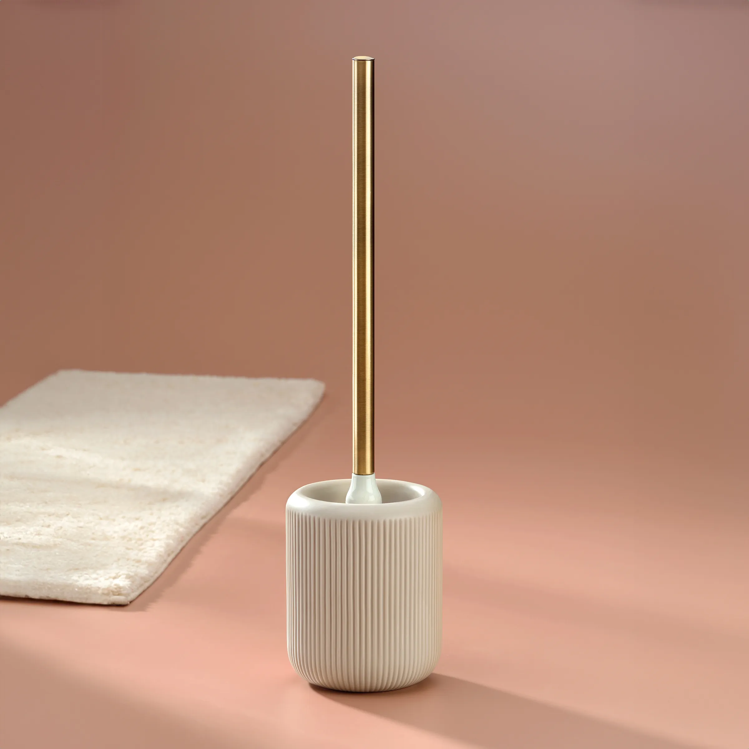 Toilettenbürste Keramik mit Gold-Griff Moderne Toilettenbürste mit weißem gerilltem Keramikbehälter und goldenem Metallstab vor rosa Hintergrund