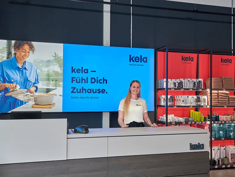Kela Heimtextilien Showroom - Feel at Home Kollektion Kela Showroom mit blauem Bildschirm, Kampagne 'Fühl Dich Zuhause', Frau an Verkaufstheke, rote Produktregale mit Heimtexilien im Hintergrund