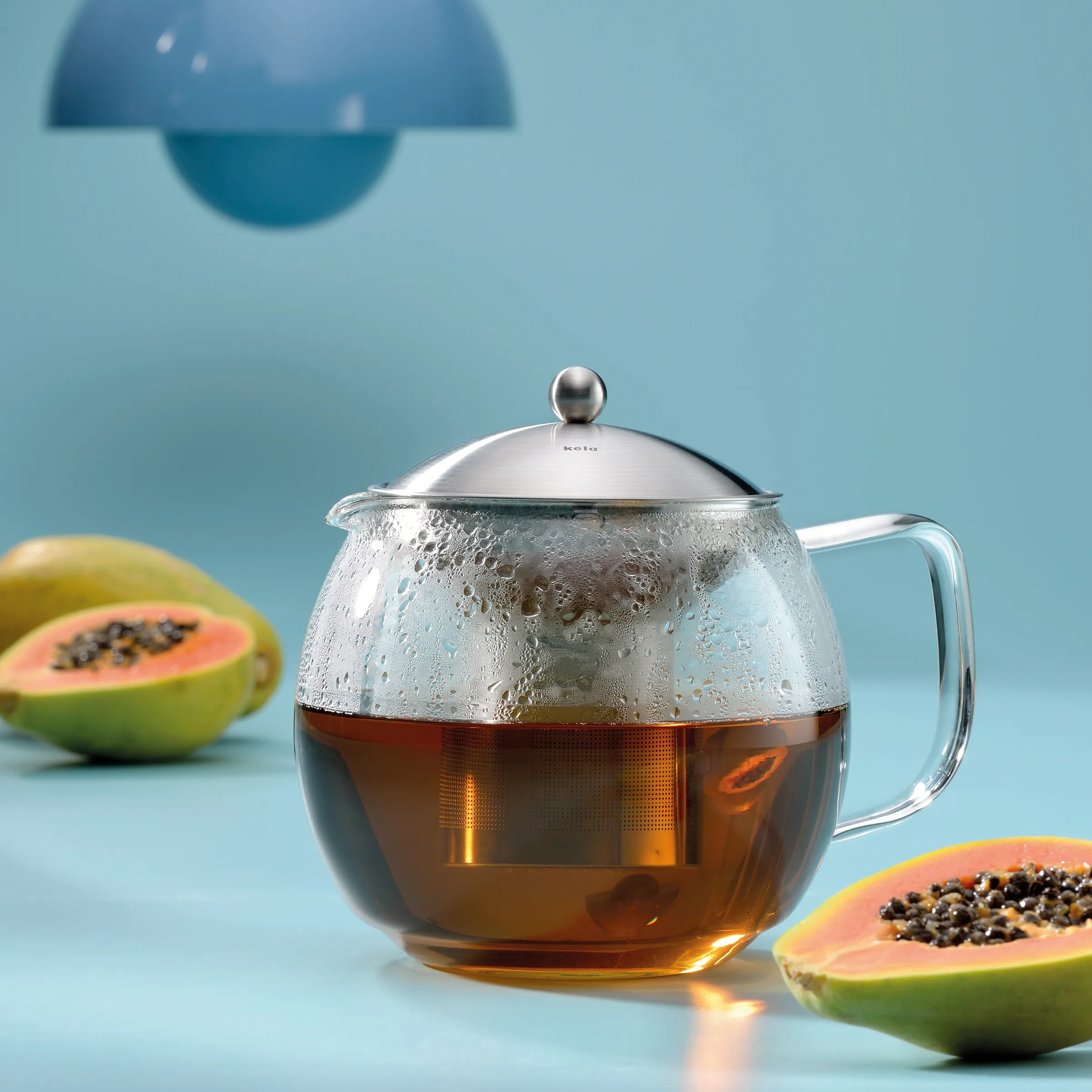 Glaskanne mit Tee und Edelstahldeckel neben halbierten Papayas auf blauem Hintergrund