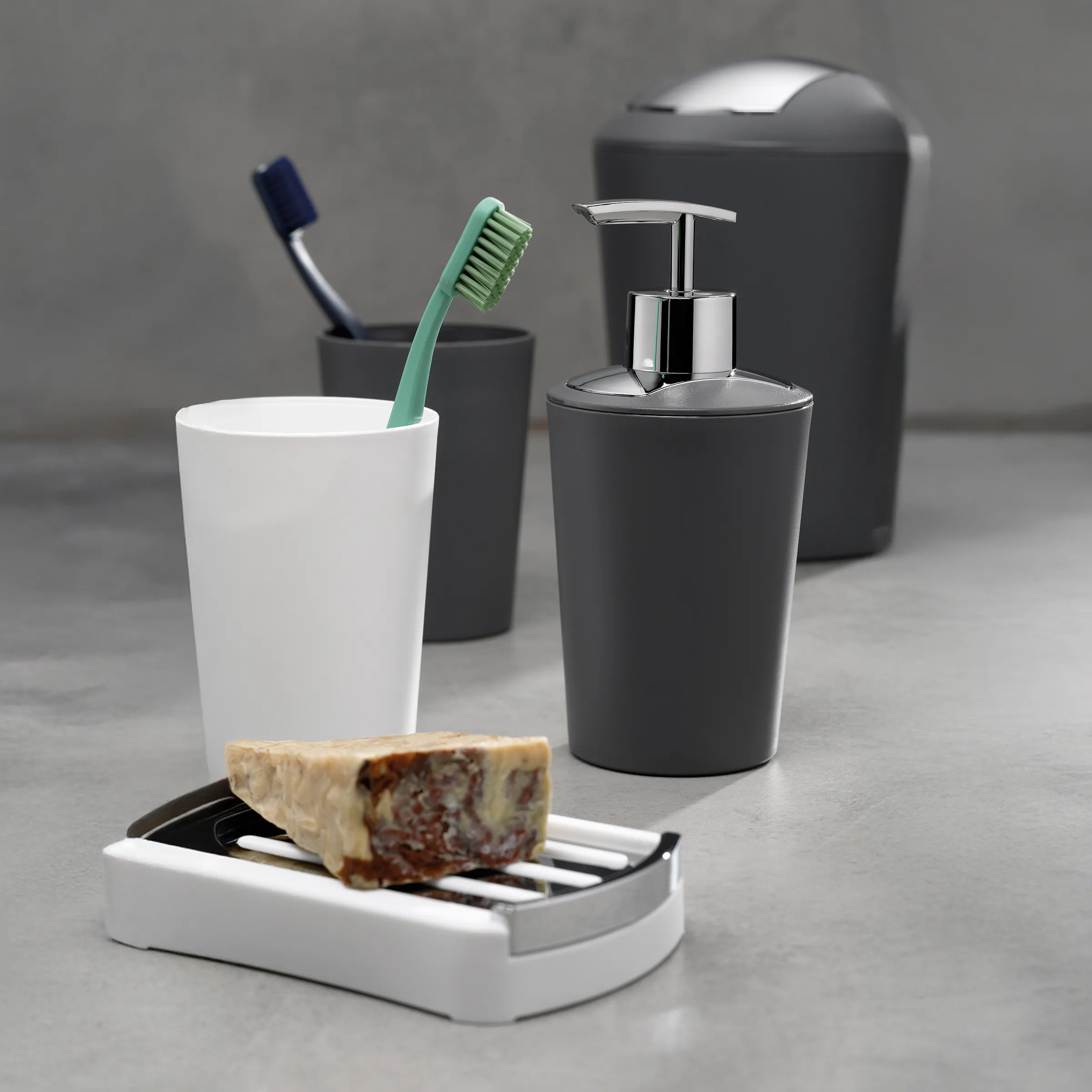 Badezimmer-Accessoires Set mit weißem Becher, grüner Zahnbürste, schwarzem Seifenspender und Behältern auf grauem Untergrund.