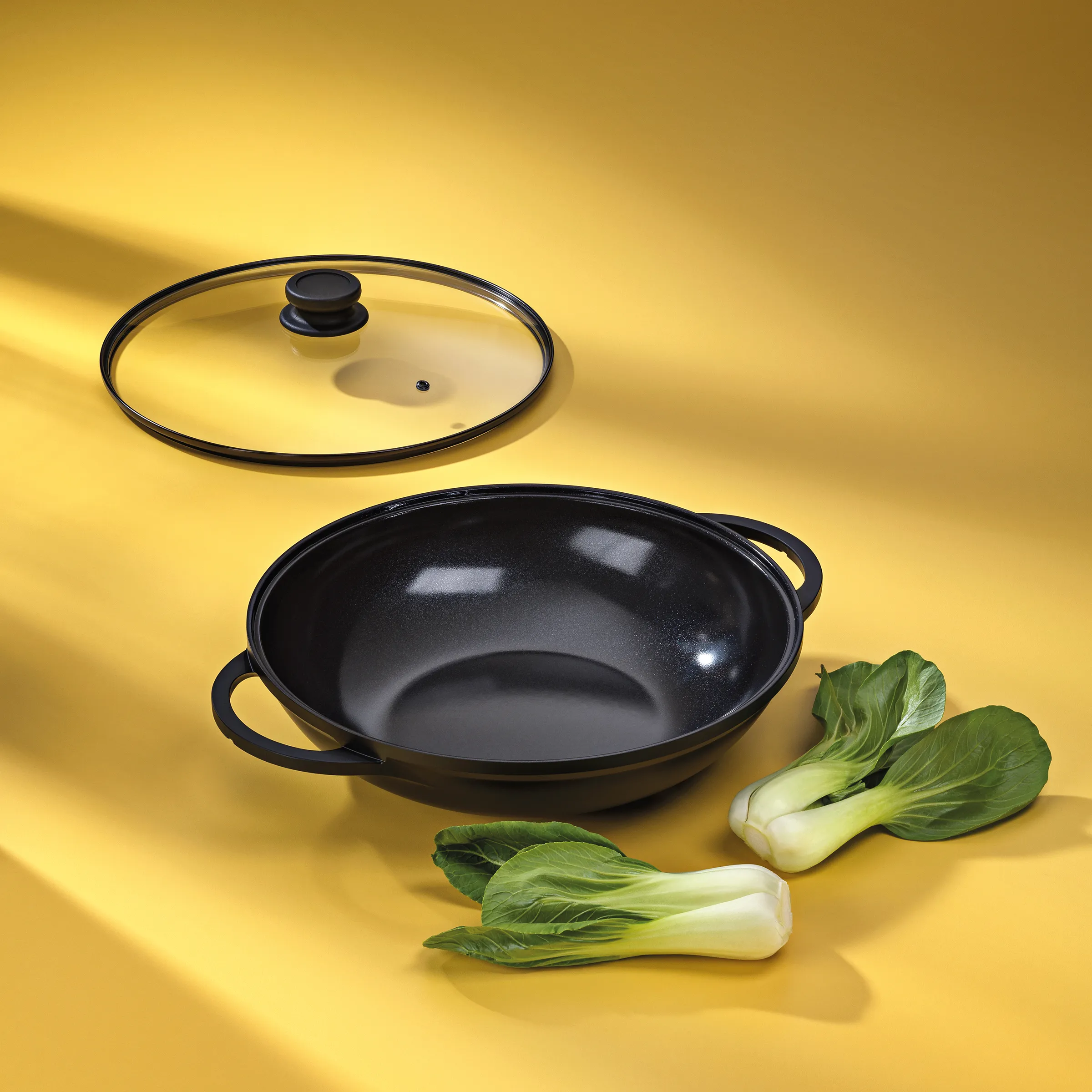 Wok mit Deckel - Asiatische Kochgeschirr Set Schwarzer Wok mit transparentem Deckel und zwei grünen Pak Choi Gemüsen auf gelbem Hintergrund