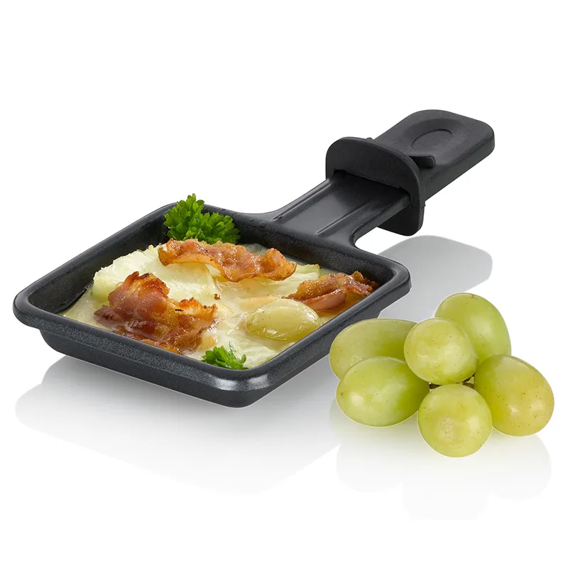 Schwarze Raclette-Pfanne mit Käse und Speck gefüllt, daneben grüne Trauben auf weißem Hintergrund
