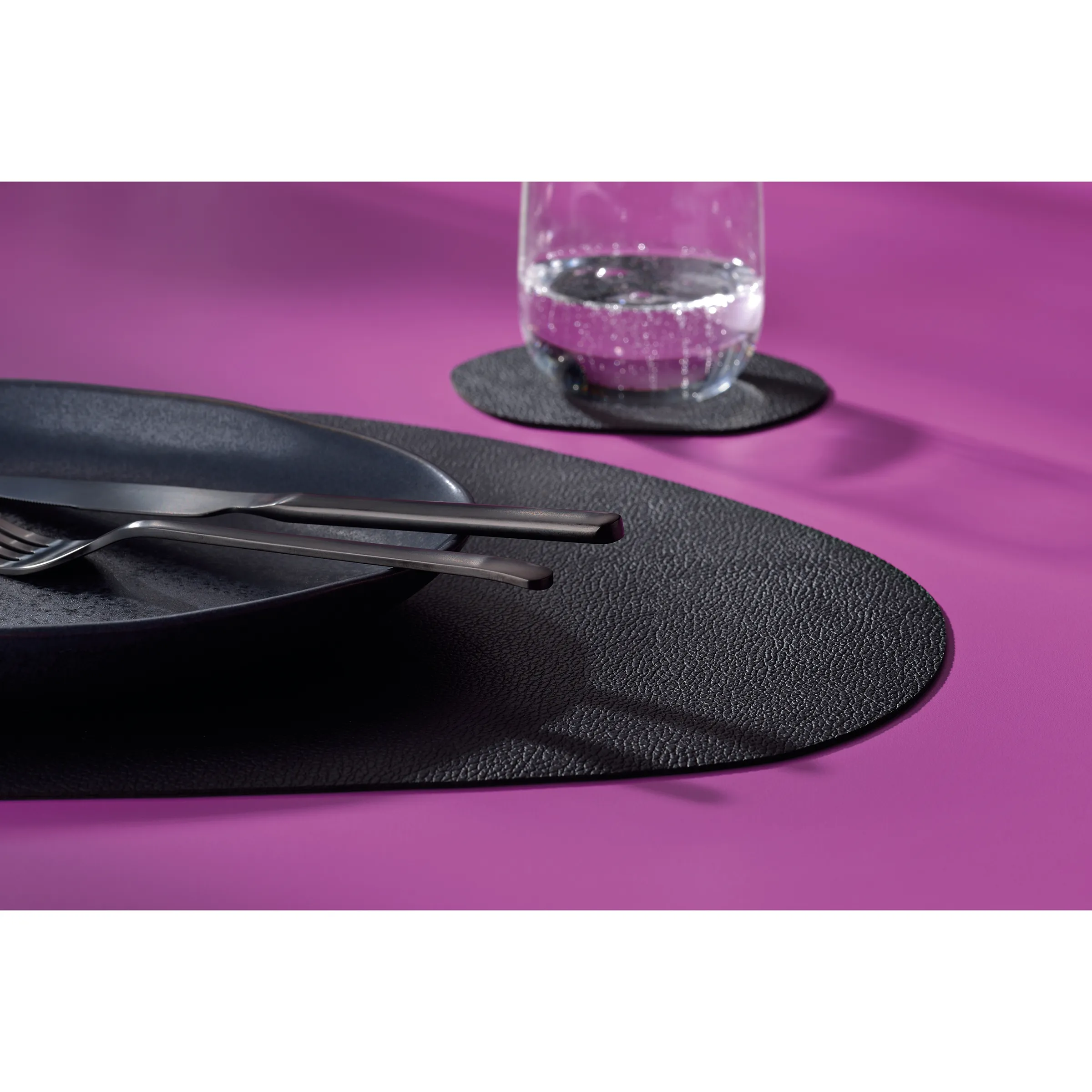 Schwarzes Leder Tischset mit Besteck und Untersetzer - Tischdekoration Schwarzes Tischset aus Leder mit Besteck und Glasuntersetzer auf violettem Hintergrund