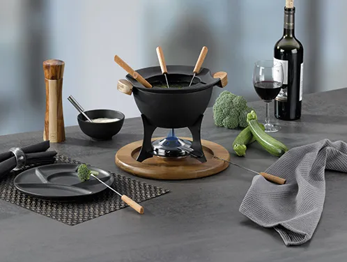 Fondue-Set schwarz mit Zubehör und Holzbesteck Schwarze Fondue-Set mit Holzbesteck, Wein, Gemüse und Zubehör auf grauem Tisch