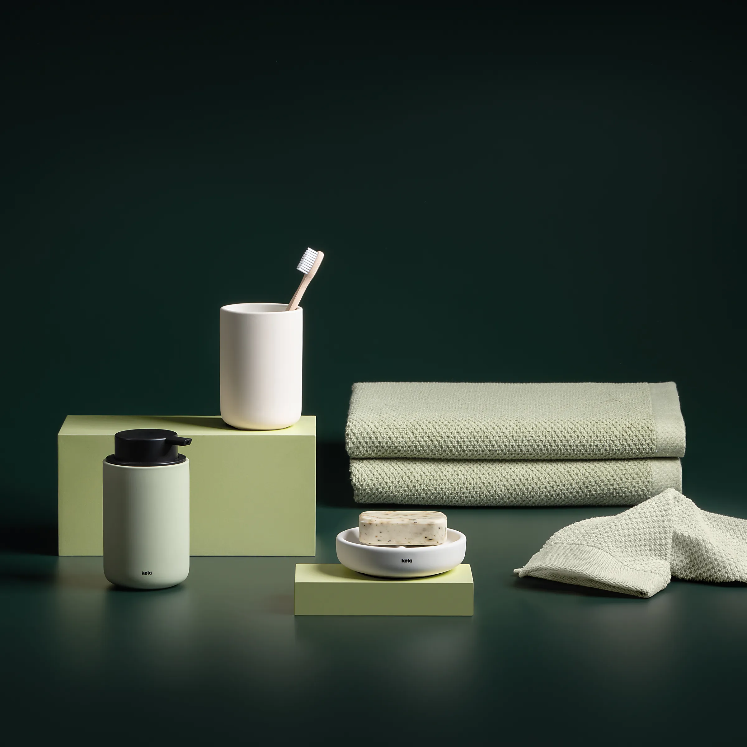 Badezimmer Accessoires Set - Design Toilettenartikel Minimalistisches Badezimmer-Set mit weißem Zahnbürstenbecher, Seifendose, beigen Handtüchern und grünen Accessoires auf dunklem Hintergrund.