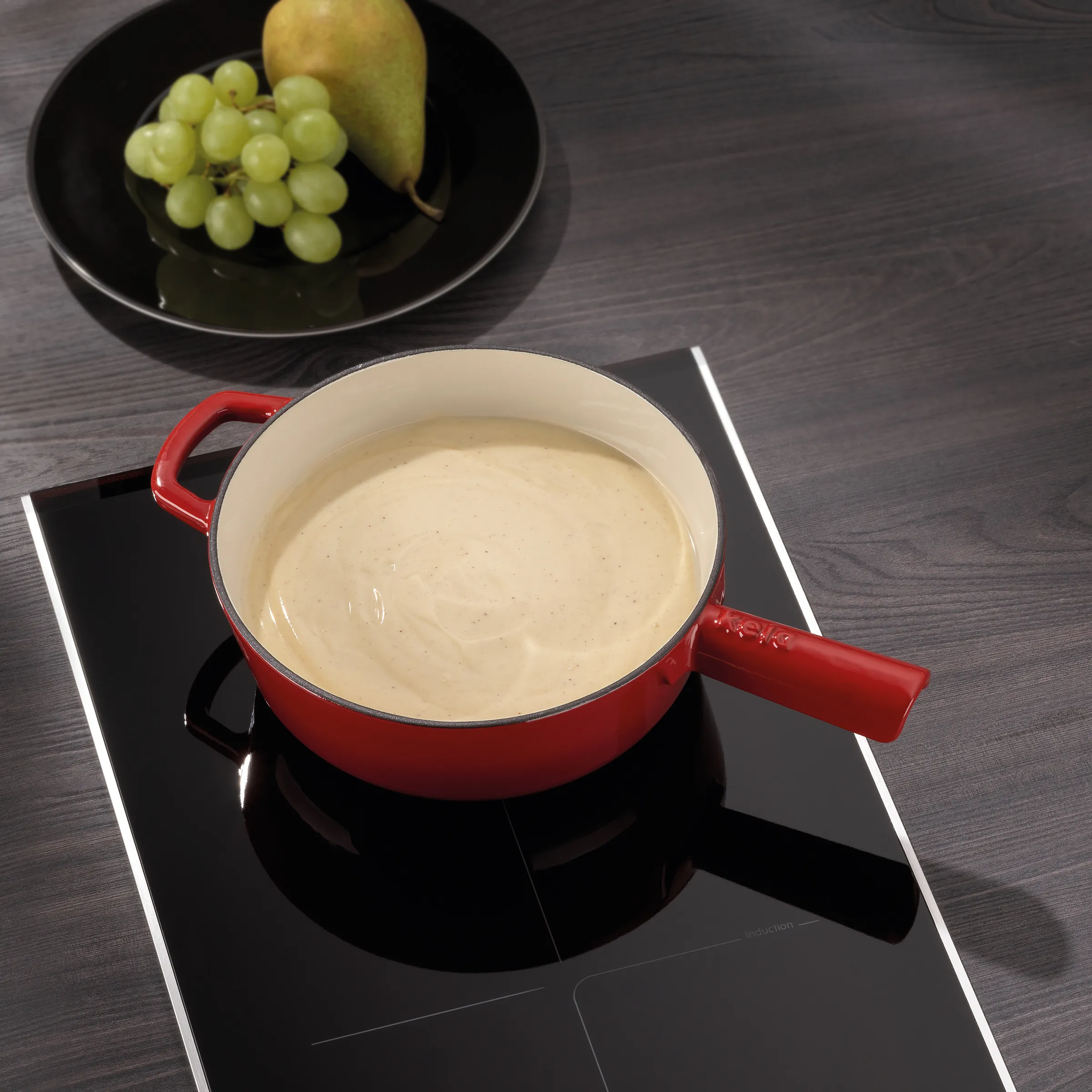 Käsefondue in roter Fonduepfanne auf Induktionsherd mit Obst Rote Fonduepfanne mit geschmolzenem Käse auf Induktionskochfeld, im Hintergrund Teller mit Trauben und Birne