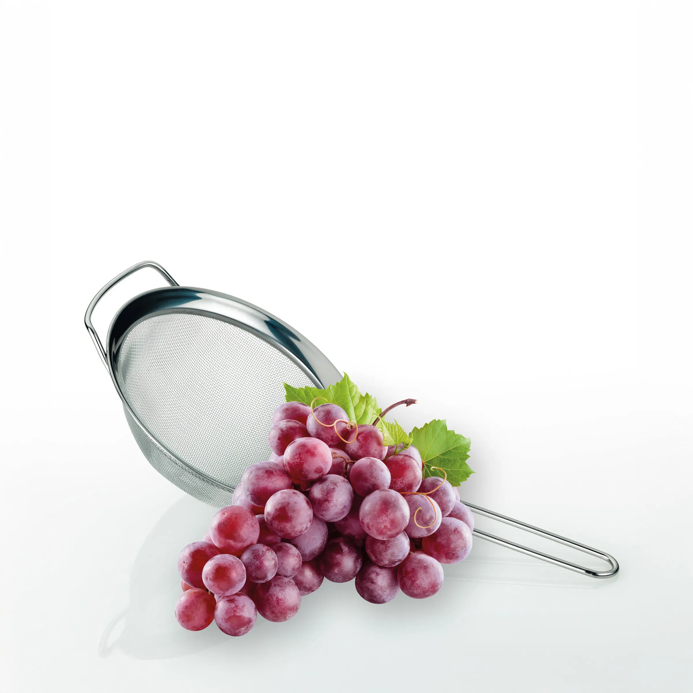 Rote Trauben Sieb - Obst waschen und servieren Rote Trauben mit grünen Blättern fallen aus einem Edelstahl-Sieb auf weißem Untergrund.