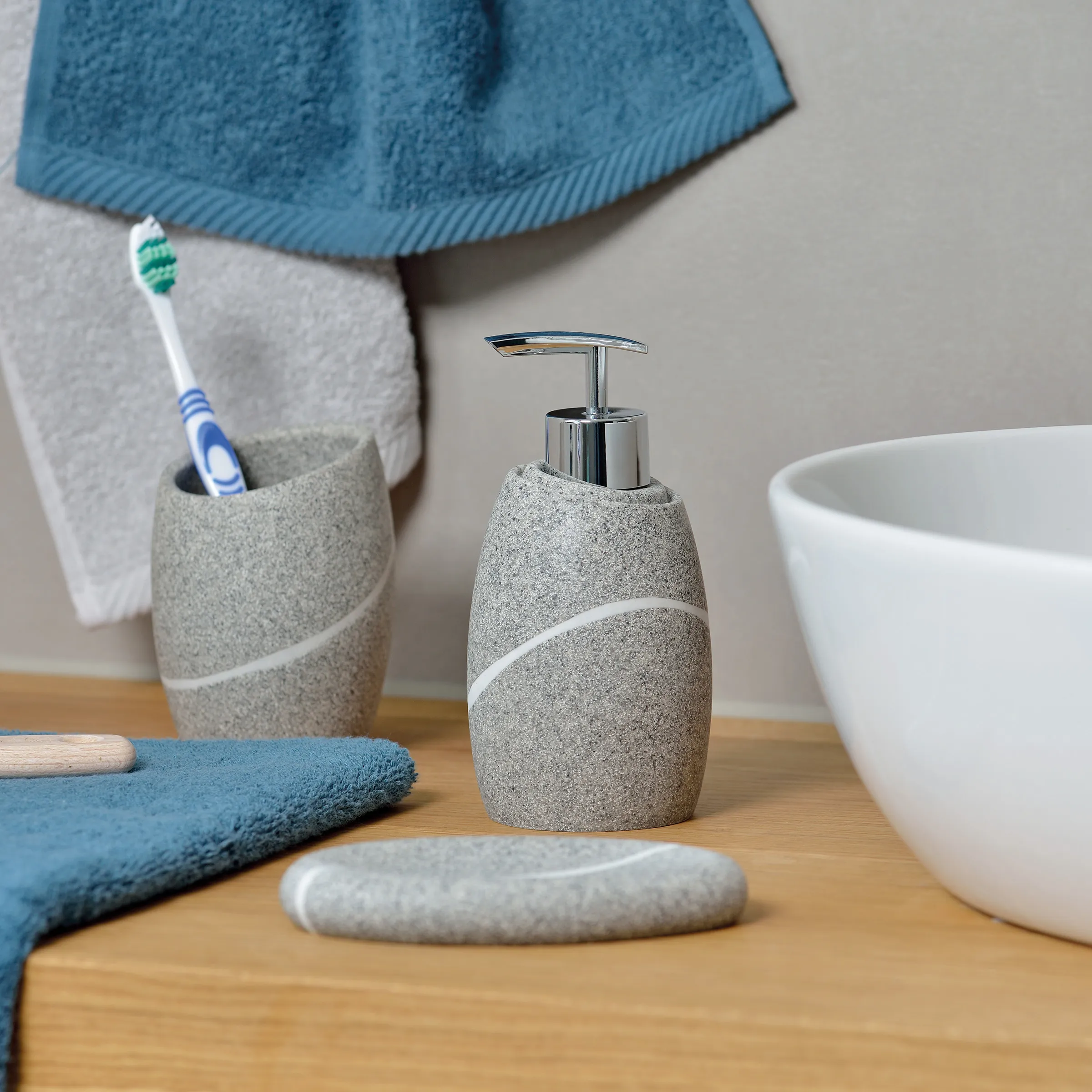 Badezimmer-Set mit grauem Seifenspender, Zahnbürstenhalter, weißer Schüssel und blauen Handtüchern auf Holztisch
