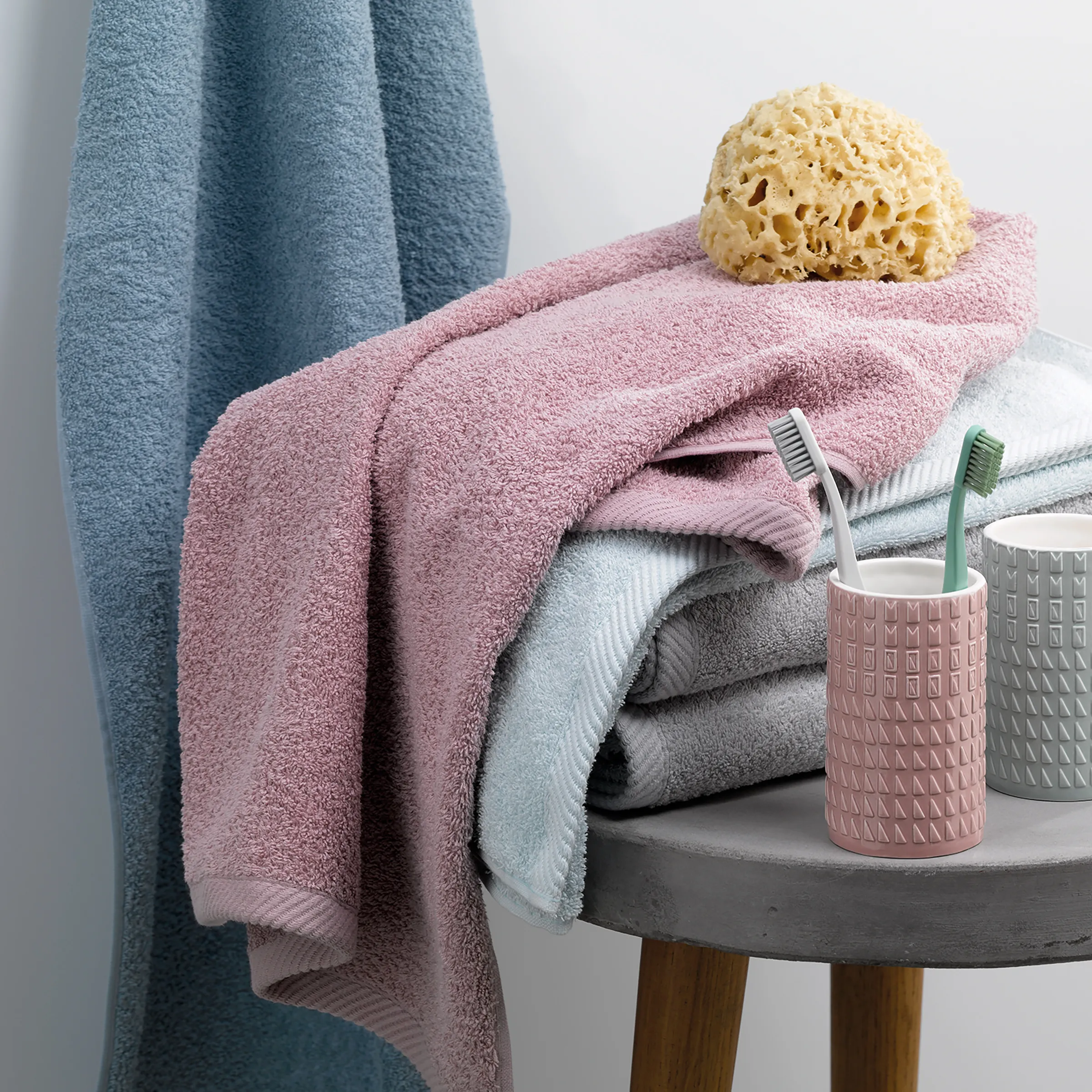 Badezimmer-Set mit Handtüchern, Badeschwamm und Accessoires Badezimmer-Arrangement mit gestapelten Handtüchern in Rosa, Blau und Weiß, Badeschwamm, Zahnbürsten und Kosmetikbehältern auf grauem Tisch