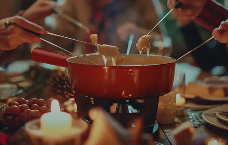 Multiple people holding fondue forks over a red fondue pot with melted cheese by candlelight during a dining experience.