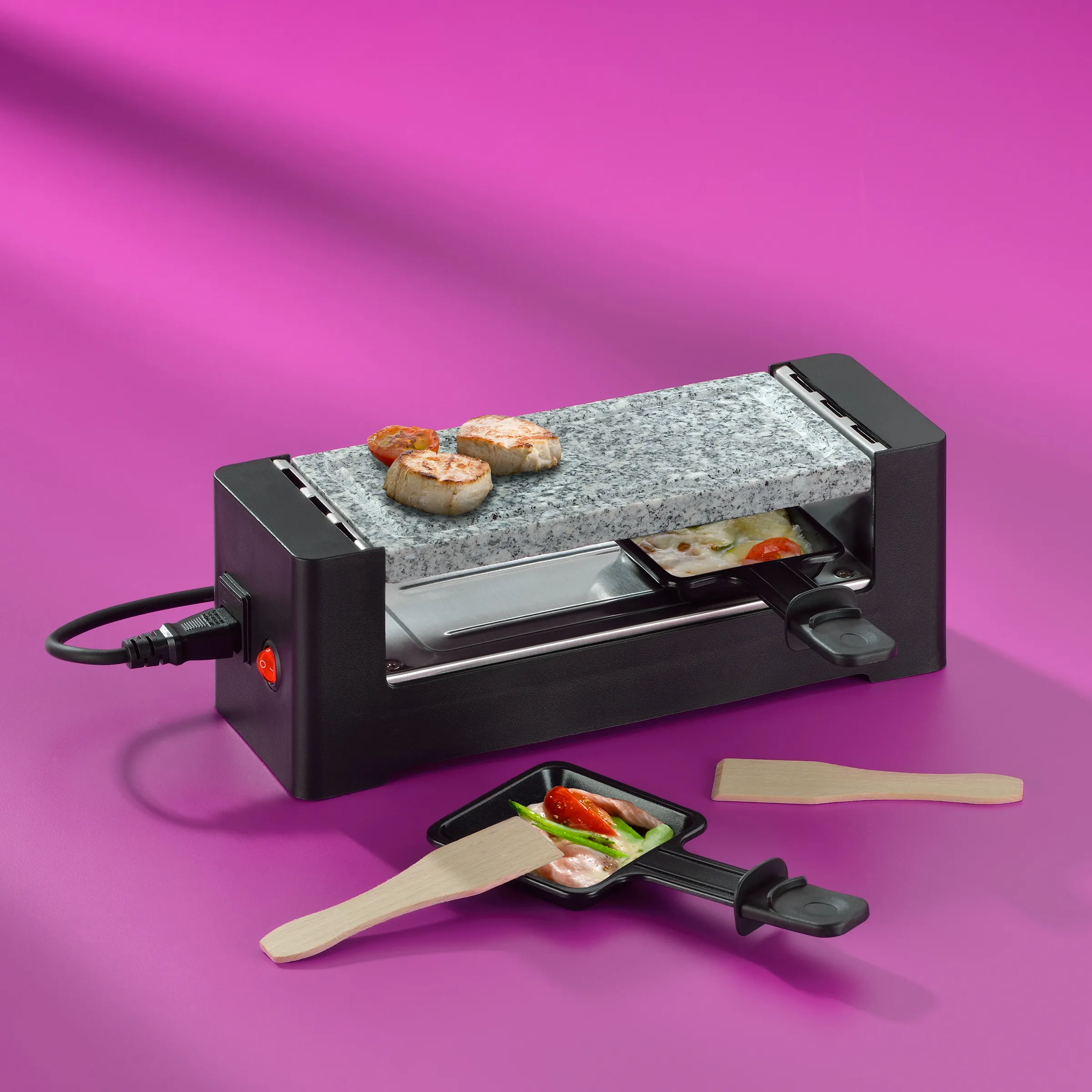 Raclette-Grill mit Granitplatte auf rosa Hintergrund, darauf gebratene Jakobsmuscheln, zwei Pfännchen mit Gemüse