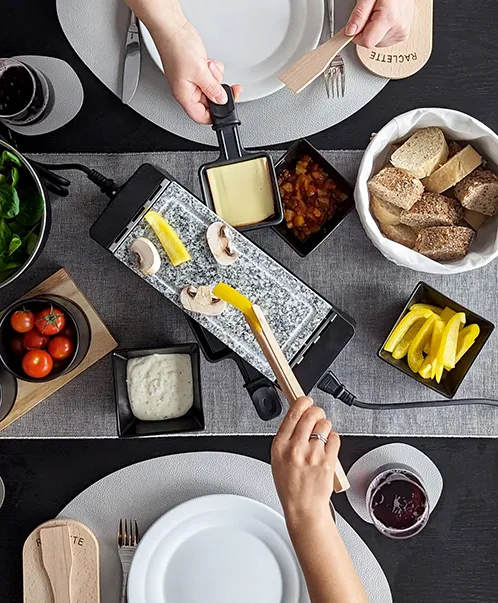 Raclette-Grill Tisch-Set mit Käse und Zutaten Raclette-Tisch-Setup mit zwei Personen, die Käse und verschiedene Zutaten auf einem Raclette-Grill zubereiten