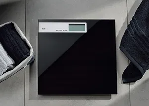 Digitale Personenwaage mit schwarzem Display und digitaler Anzeige auf grauem Fliesenboden, umgeben von weißen Handtüchern