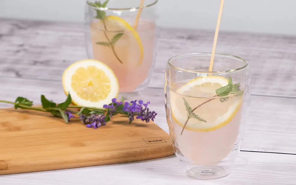 Erfrischende Limonade mit Zitrone und Lavendel Zwei Gläser mit hellrosa Getränk, Zitronenscheiben, Minze und Strohhalmen auf Holzbrett neben Lavendelblüten