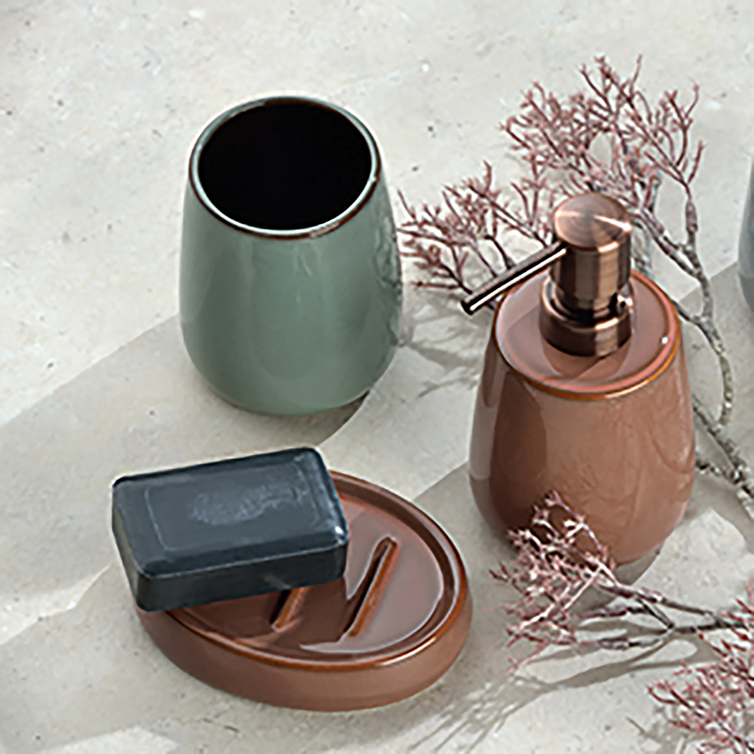 Badezimmer-Accessoires Set: Zahnputzbecher & Seifenspender Badezimmer-Set bestehend aus grüner Zahnputzbecher, brauner Seifenschale mit schwarzer Seife und brauner Seifenspender mit getrockneter Pflanze auf hellem Untergrund.