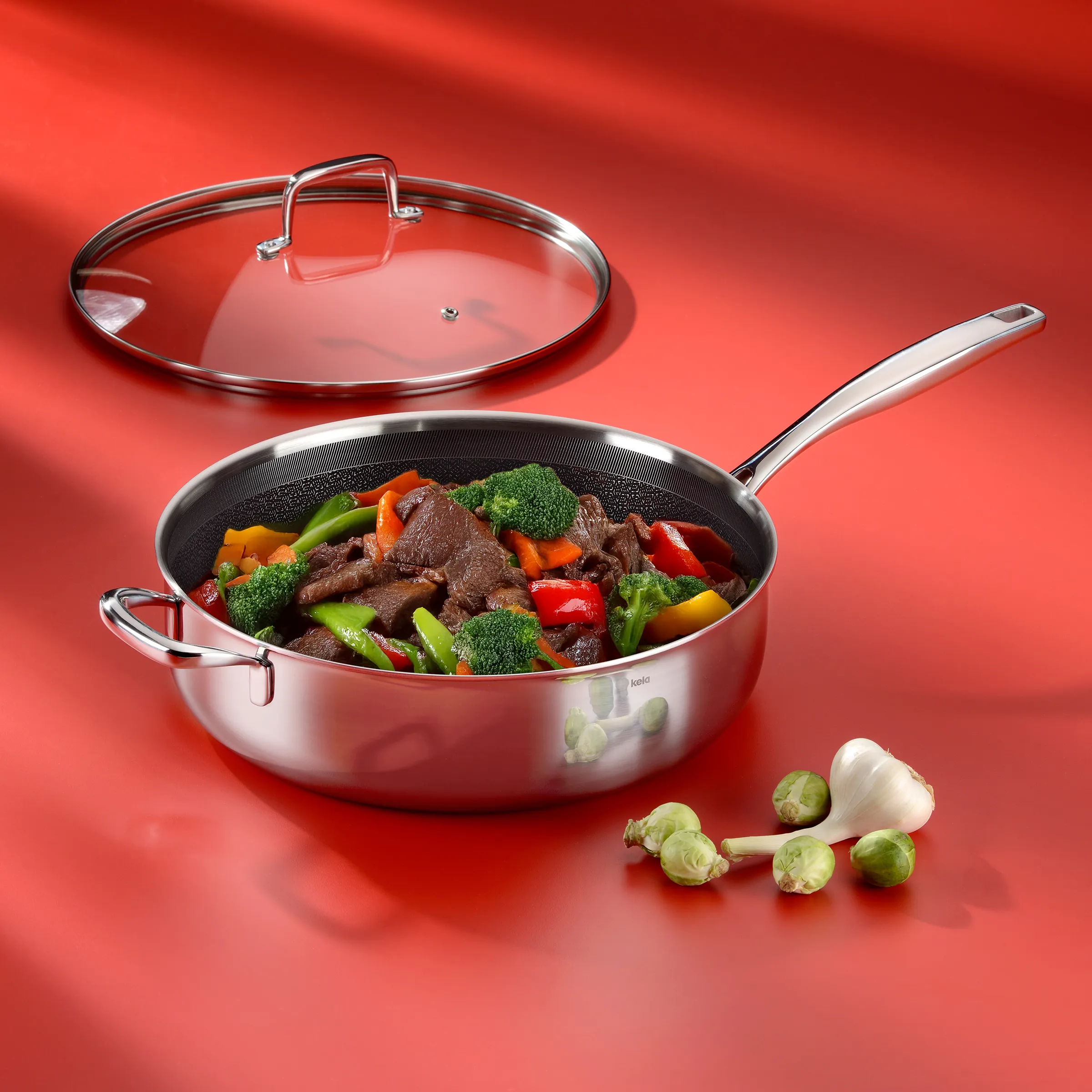Edelstahl-Sauteuse mit Glasdeckel - Bratpfanne für Fleisch und Gemüse Edelstahl-Sauteuse mit Glasdeckel, gefüllt mit Rindfleisch und buntem Gemüse auf rotem Hintergrund