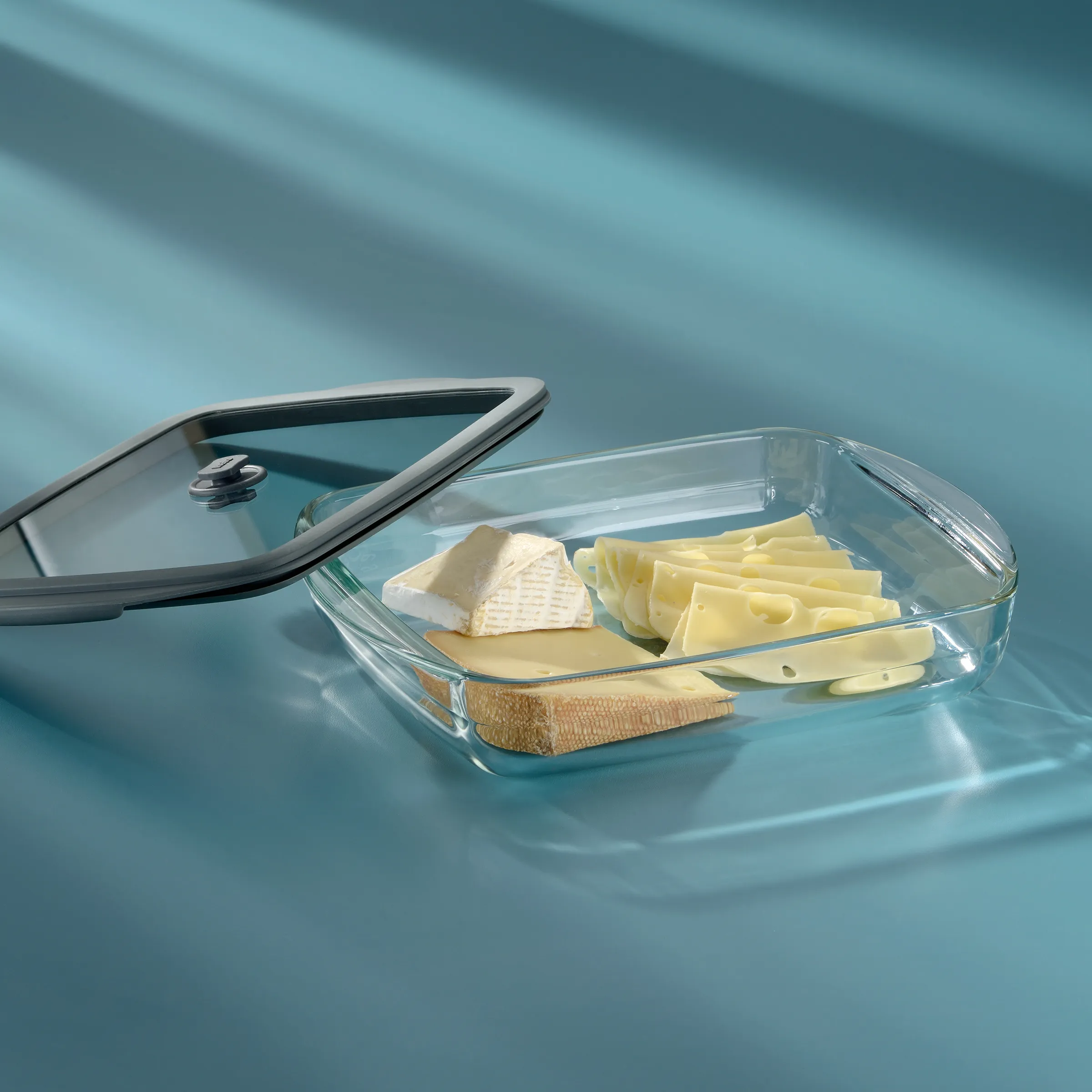 Glasauflaufform mit Käse, Butter und Brot auf blauem Hintergrund mit Kochlöffel