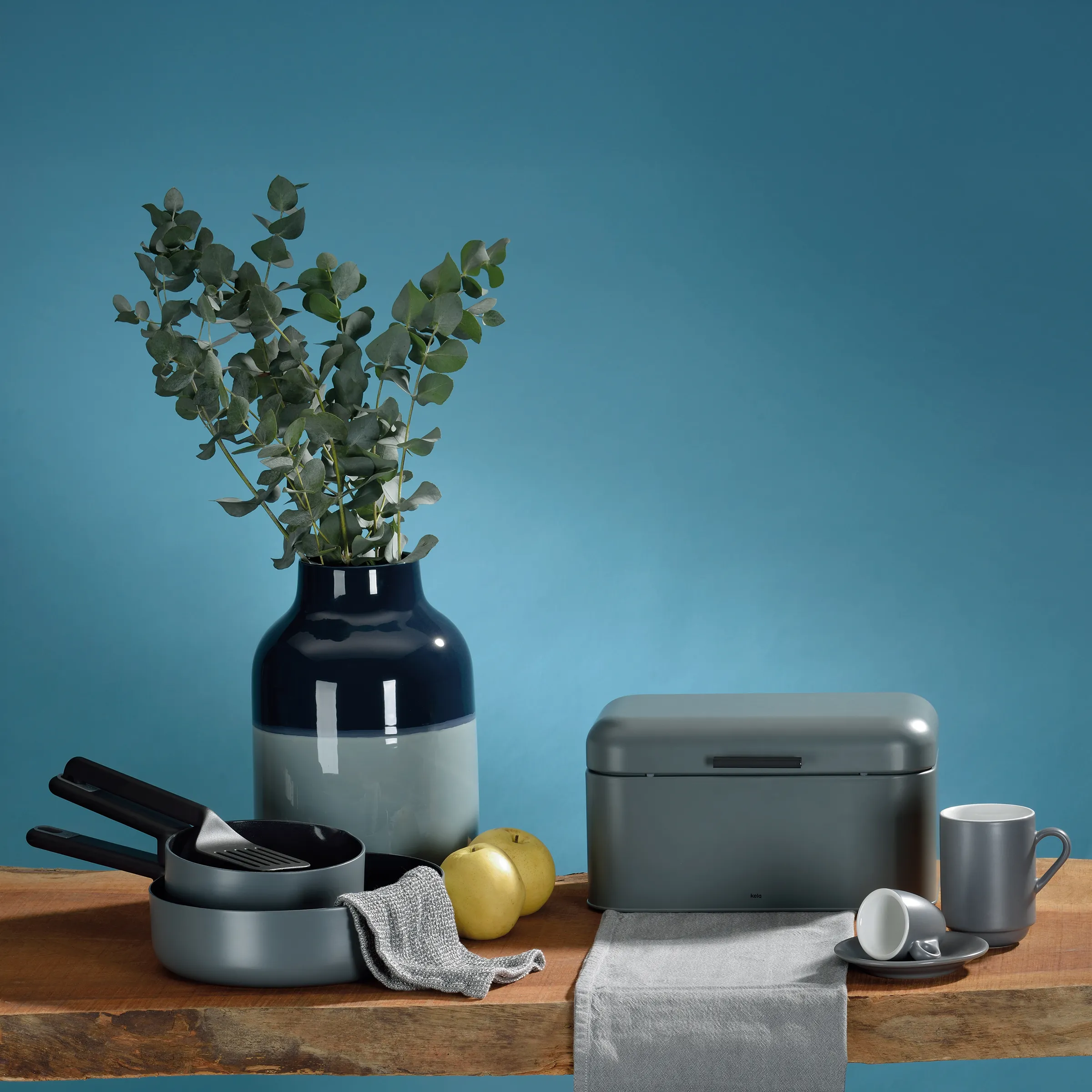 Moderne Küchenausstattung und Küchendekor Set Küchenausstattung auf Holztisch: schwarze Vase mit Eukalyptuszweigen, graue Töpfe, Besteck, gelbe Äpfel, graue Mug und Aufbewahrungsbox vor blauem Hintergrund