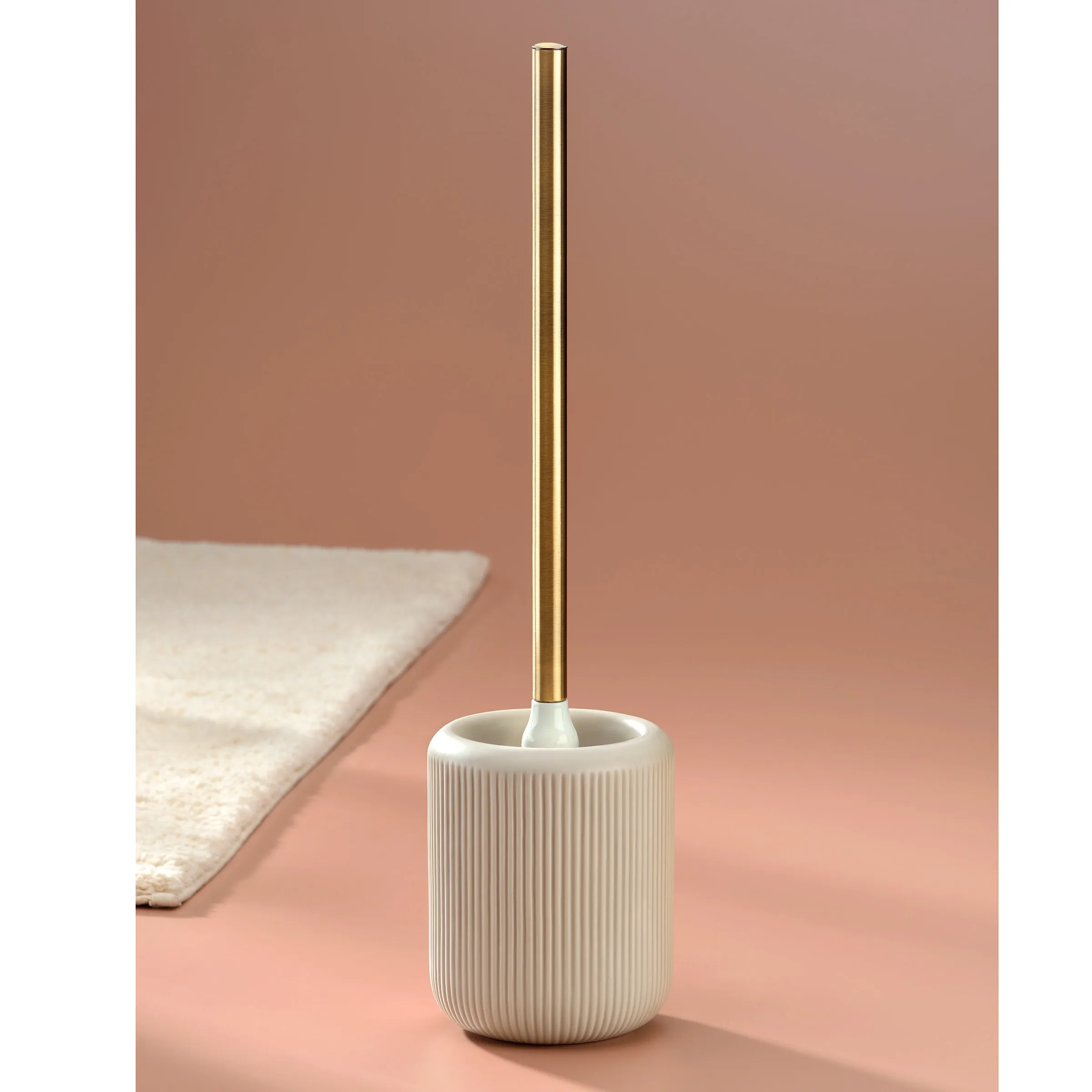 Toilettenbürste mit Keramikhalter und Goldgriff Weiße Toilettenbürste mit geripptem Keramikbehälter und goldenem Metallstab auf rosa Hintergrund neben Handtuch