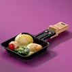 Schwarze Raclettepfanne mit geschmolzenem Käse, Kartoffeln und Gemüse auf violettem Hintergrund