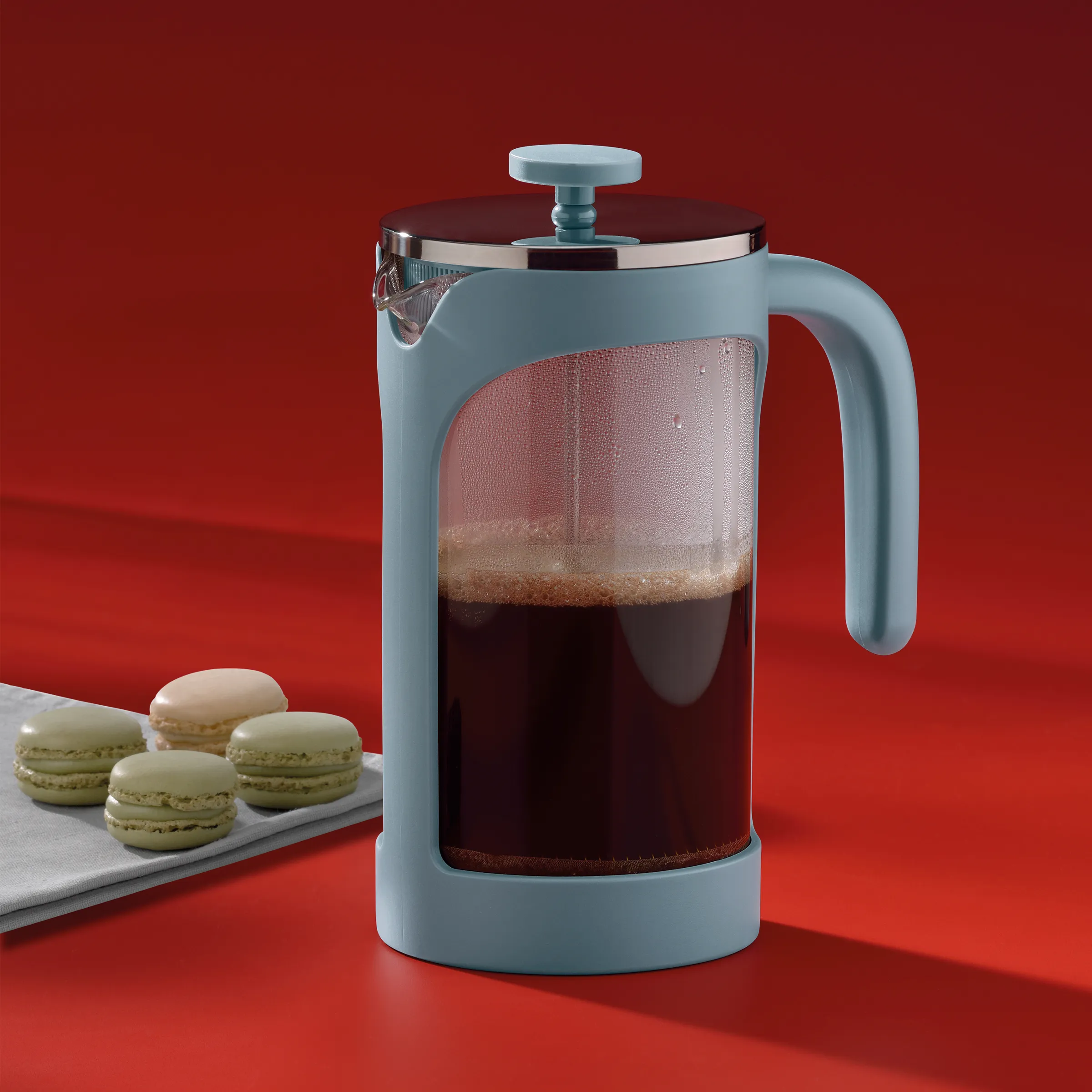 Pressefilter Kaffeepresse blau für frischen Kaffee Hellblauer Pressefilter mit schwarzem Deckel gefüllt mit dunkelbraunem Kaffee, daneben Macarons auf weißem Tablett vor rotem Hintergrund