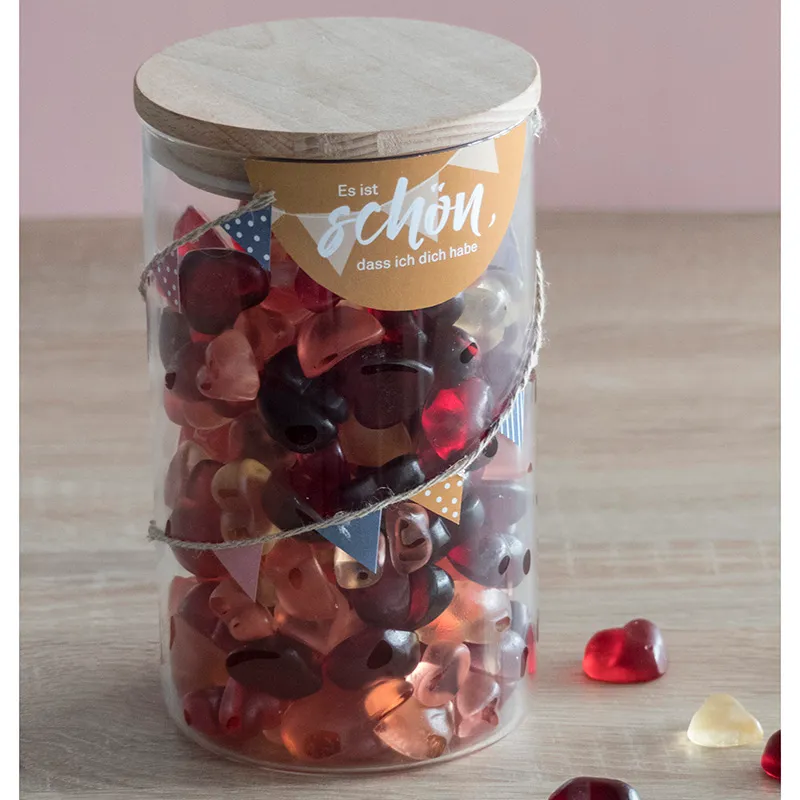 Glasdose mit Holzdeckel gefüllt mit roten Fruchtgummis, dekoriert mit Banner und Etikett 'Es ist schön, dass ich dich habe'