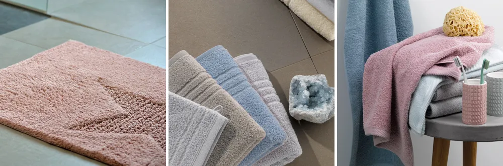 Kollektion von Badtextilien: rosa Badematte, bunte Handtücher in Grau, Blau und Beige, sowie dekorative Accessoires auf Holzregal.