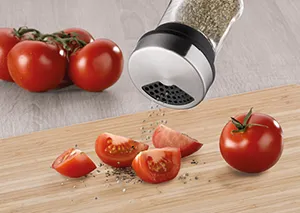 Pfeffermühle mit Tomaten - Gewürzmühle für frisch gemahlenen Pfeffer Pfeffermühle streut schwarzen Pfeffer über geschnittene Tomaten auf Holzbrett