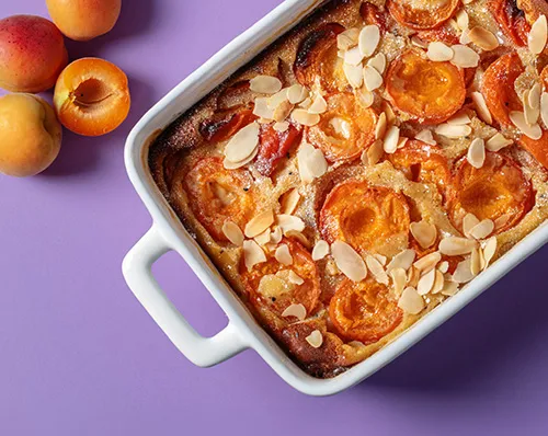 Aprikosen-Mandel-Kuchen in weißer Auflaufform mit frischen Aprikosen auf violettem Hintergrund
