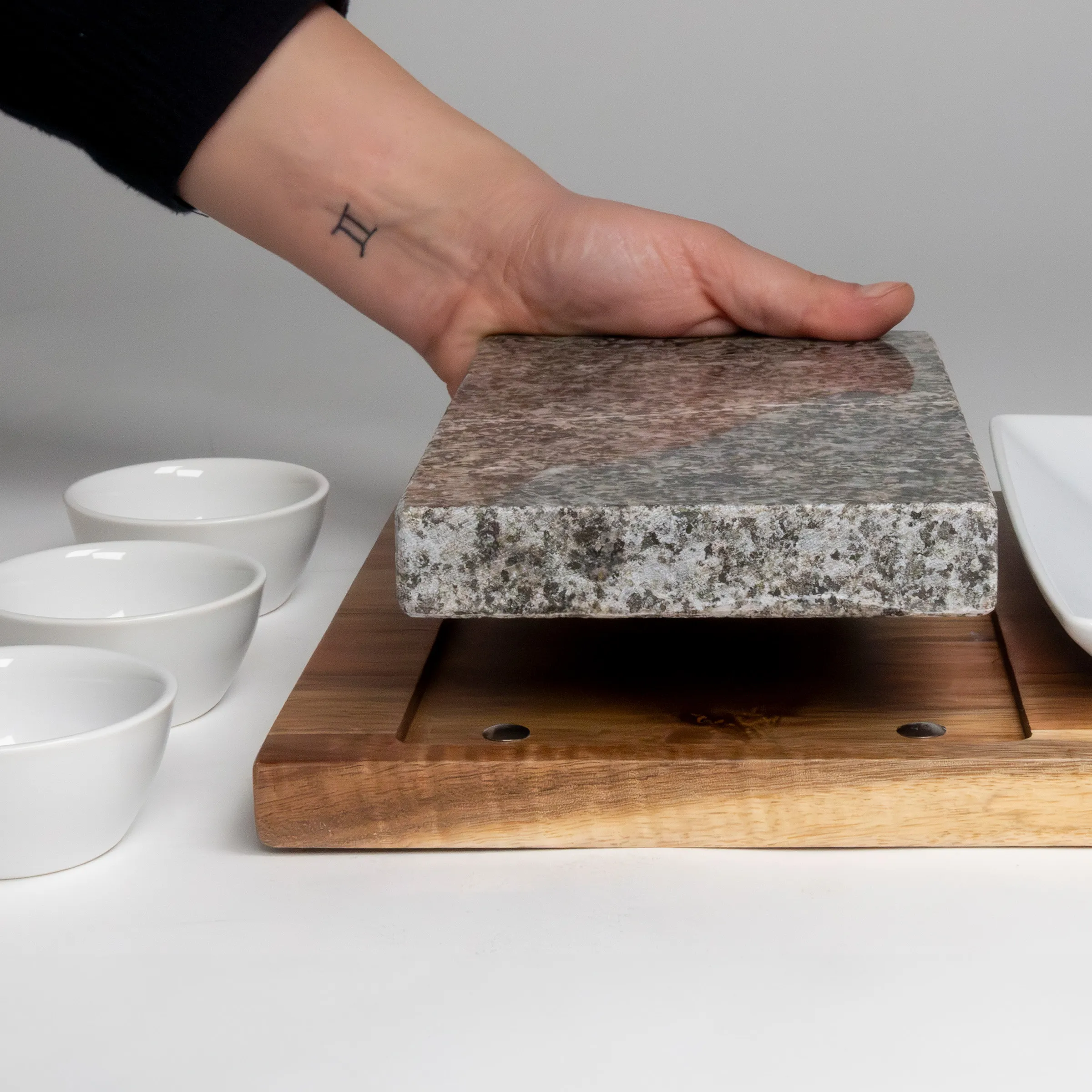 Grillstein aus Granit auf Holzbrett - Hot Stone Grill Set mit Servierschalen Hand berührt Grillstein auf Holzbrett neben weißen Schalen, Zwillinge-Tattoo am Handgelenk
