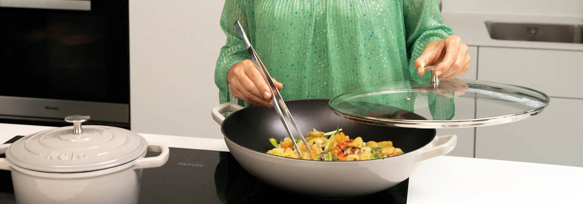 Wok mit Gemüse - Asiatisches Kochen in der Küche Person in grüner Jacke rührt bunte Gemüsepfanne in Wok mit Metalldeckel in moderner Küche