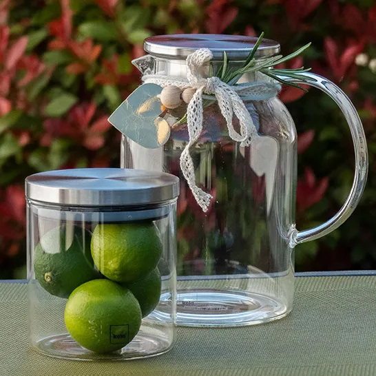 Carafe en Verre et Verre Limonade Citrons Verts pour Distributeur Carafe en verre et verre avec citrons verts et herbes aromatiques, décorés d'un ruban crème devant un jardin coloré