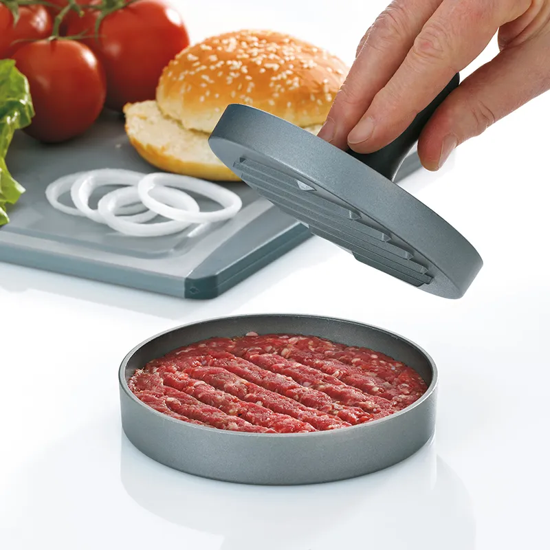 Burgerpresse für perfekte Hamburger Patties - Hackfleisch Presse Hand drückt Burgerpresse mit Hackfleisch-Patty, Hamburgerbrötchen und Tomatenscheiben im Hintergrund