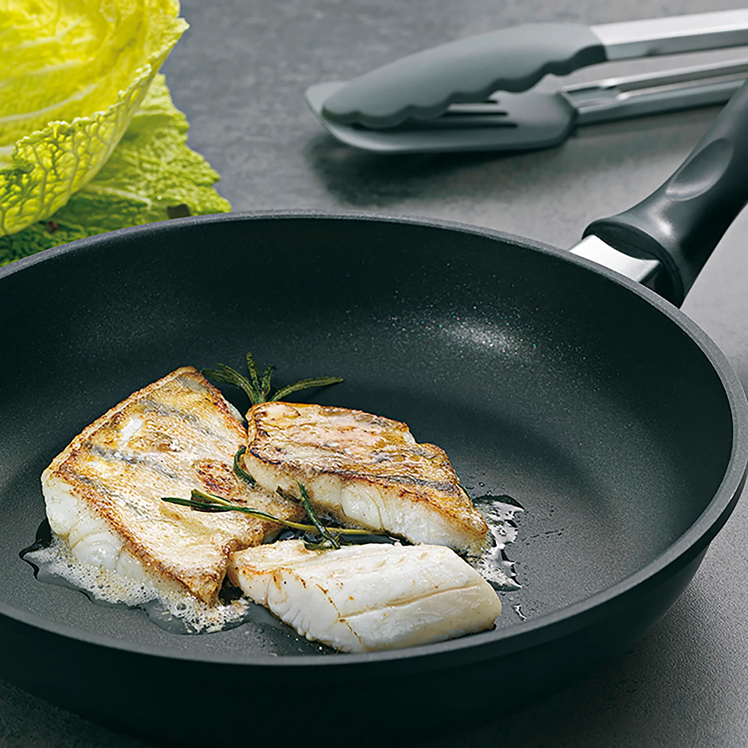 Fischfilets braten - Premium Kochzubehör und Küchenwerkzeuge Gebratene Fischfilets in schwarzer Pfanne mit Rosmarin und frischem Gemüse auf grauem Untergrund