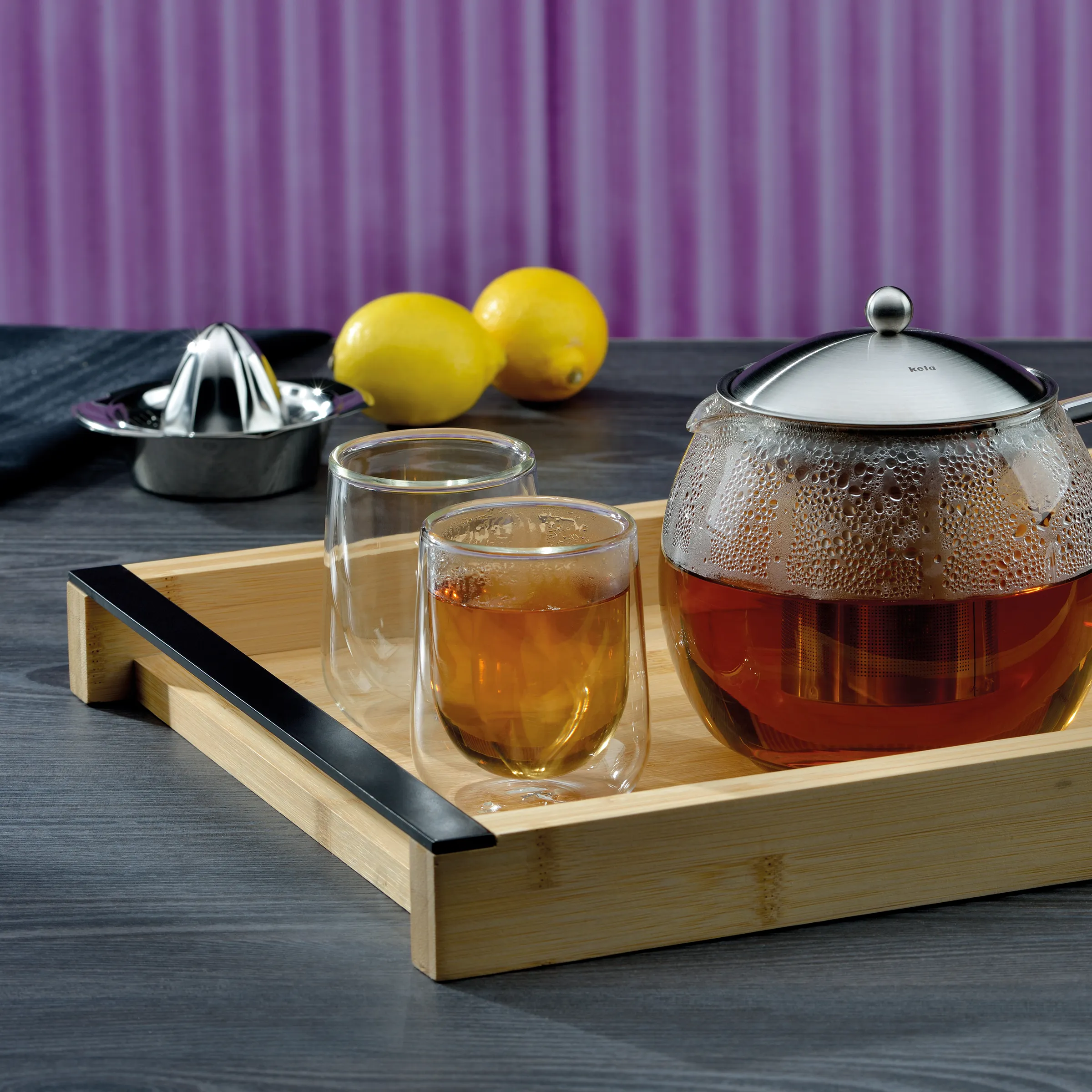 Teeset Glas mit Kanne und Tassen - Teeservice Teeset mit Glaskanne gefüllt mit Tee, zwei Teegläsern auf Holztablett, Zitronen und Zitronenpresse im Hintergrund vor lila Vorhang