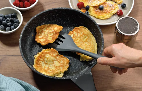 Kartoffelpuffer braten - Rezept mit frischen Beeren Hand wendet Pfannenwender bei Kartoffelpuffern in schwarzer Pfanne an, umgeben von Beeren und Toppings.