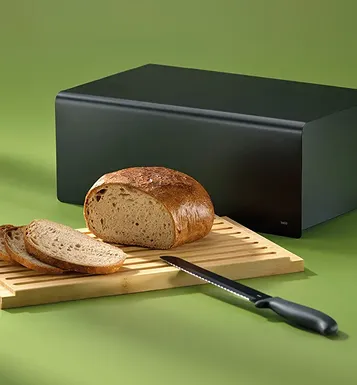Schwarzer Brotkasten neben aufgeschnittenem Vollkornbrot auf Holzbrett mit Messer auf grünem Hintergrund