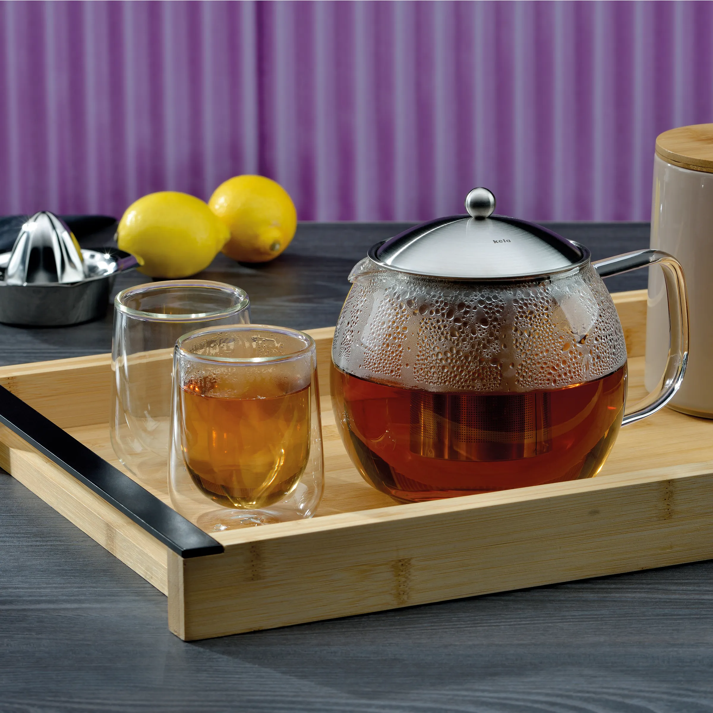Glas-Teekanne Set mit doppelwandigen Teegläsern auf Bambustablett Glas-Teekanne mit Tee und zwei doppelwandigen Gläsern auf Bambustablett, Zitronen und Zitruspresse im Hintergrund