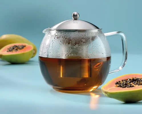 Glas-Teekanne mit Tee und frischer Papaya - Teezubereitung Glas-Teekanne mit aufgebrühtem Tee und Edelstahldeckel, daneben aufgeschnittene Papayas auf blauem Hintergrund
