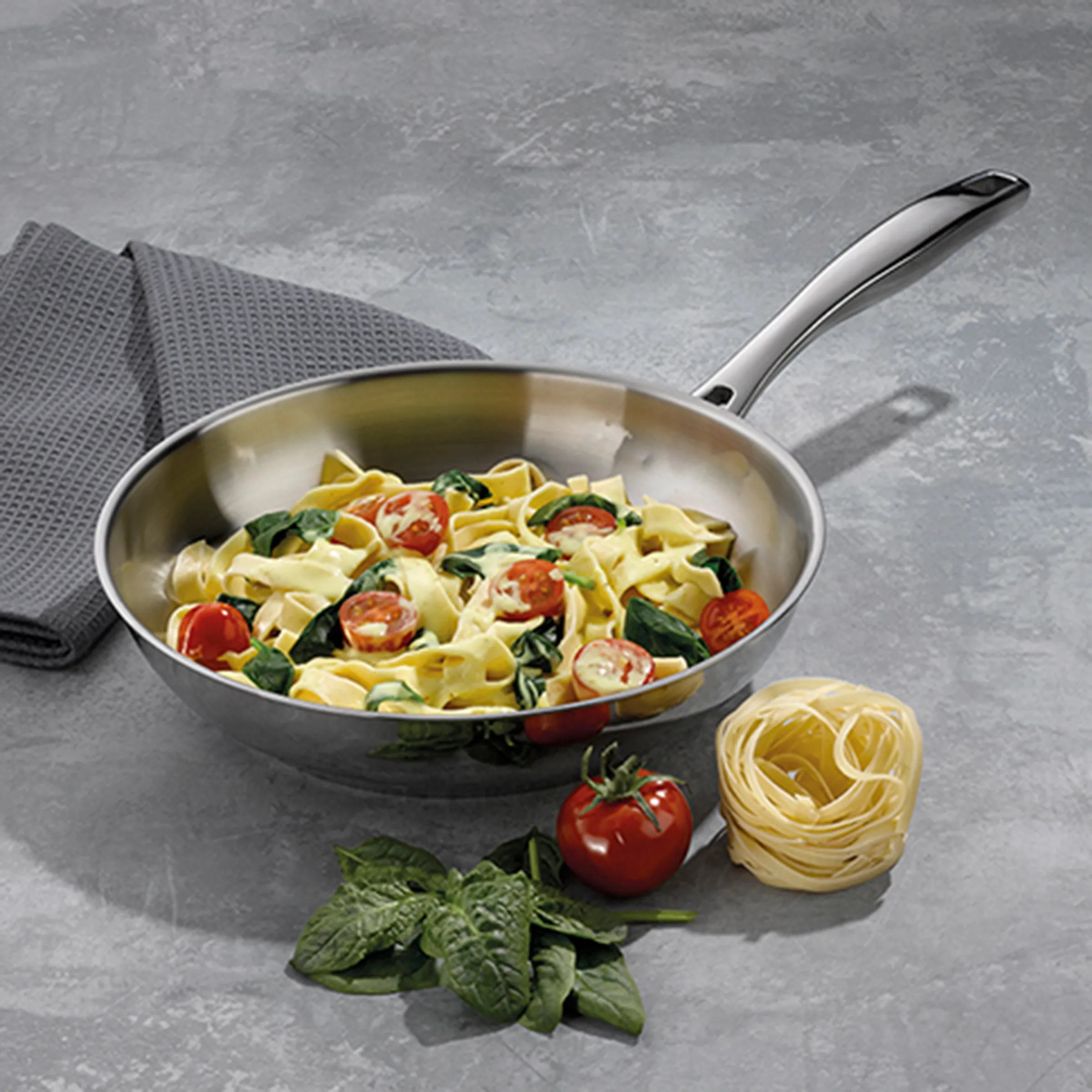 Pasta Pfanne mit Gemüse - Edelstahl Kochgeschirr Edelstahl-Pfanne mit Pasta, Tomaten und Spinat auf grauem Untergrund mit frischen Zutaten