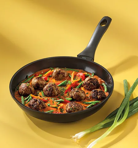 Schwarze Pfanne mit Fleischbällchen, Paprika und grünen Bohnen auf gelbem Hintergrund mit Lauch