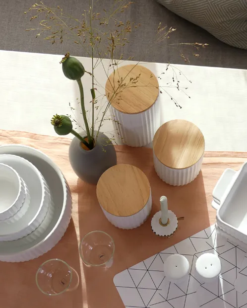 Moderne Tischdeko mit Holzbehältern und Porzellanbesteck Tischgedeck mit weißem Porzellan, Holzbehältern mit Deckeln und Blumenvase mit grünen Knospen auf Holztablett.